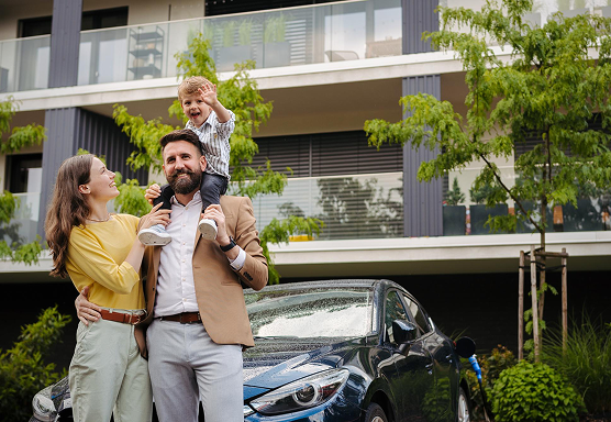 Lächelnde Familie vor einer Eigentumswohnung, die grün strahlt. Ebenfalls steht ein Auto in der Einfahrt.