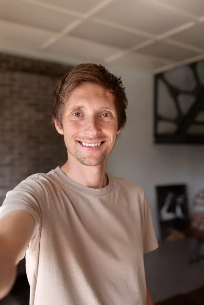 Smiling man with short brown hair wearing a beige t-shirt taking a selfie indoors.