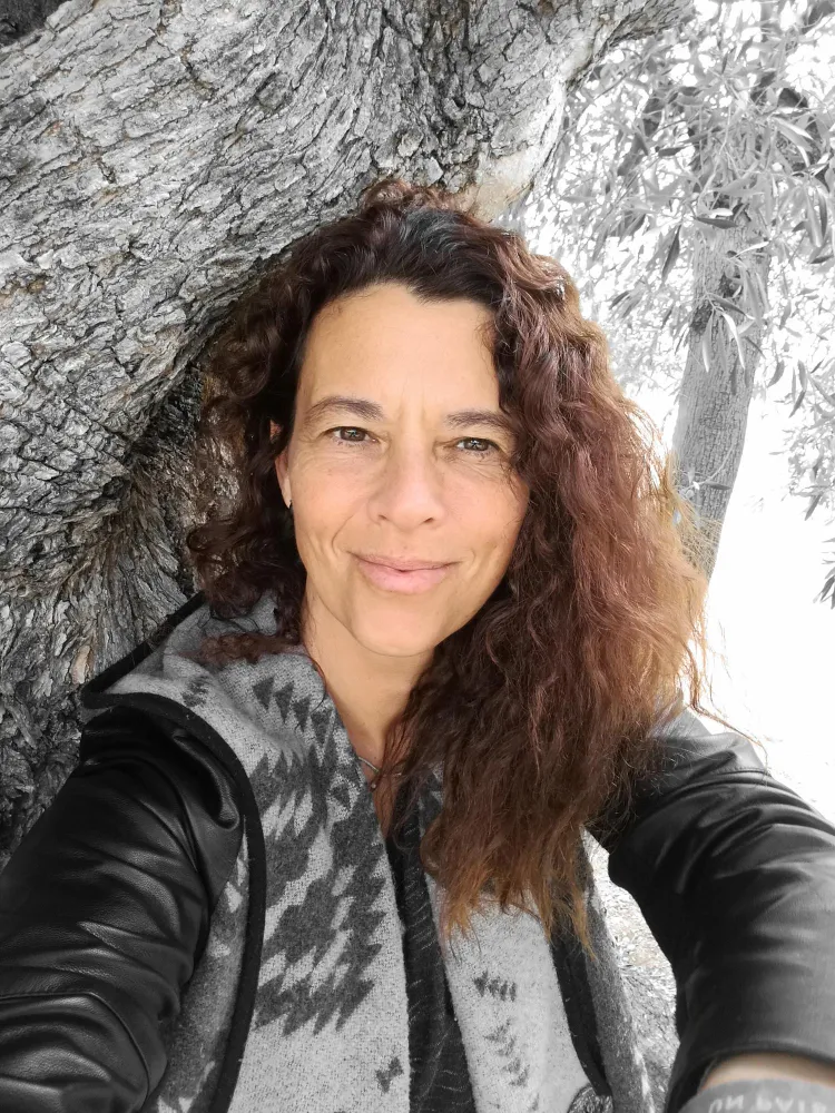 Woman with curly brown hair wearing a patterned gray and black jacket, smiling in front of a large tree trunk.