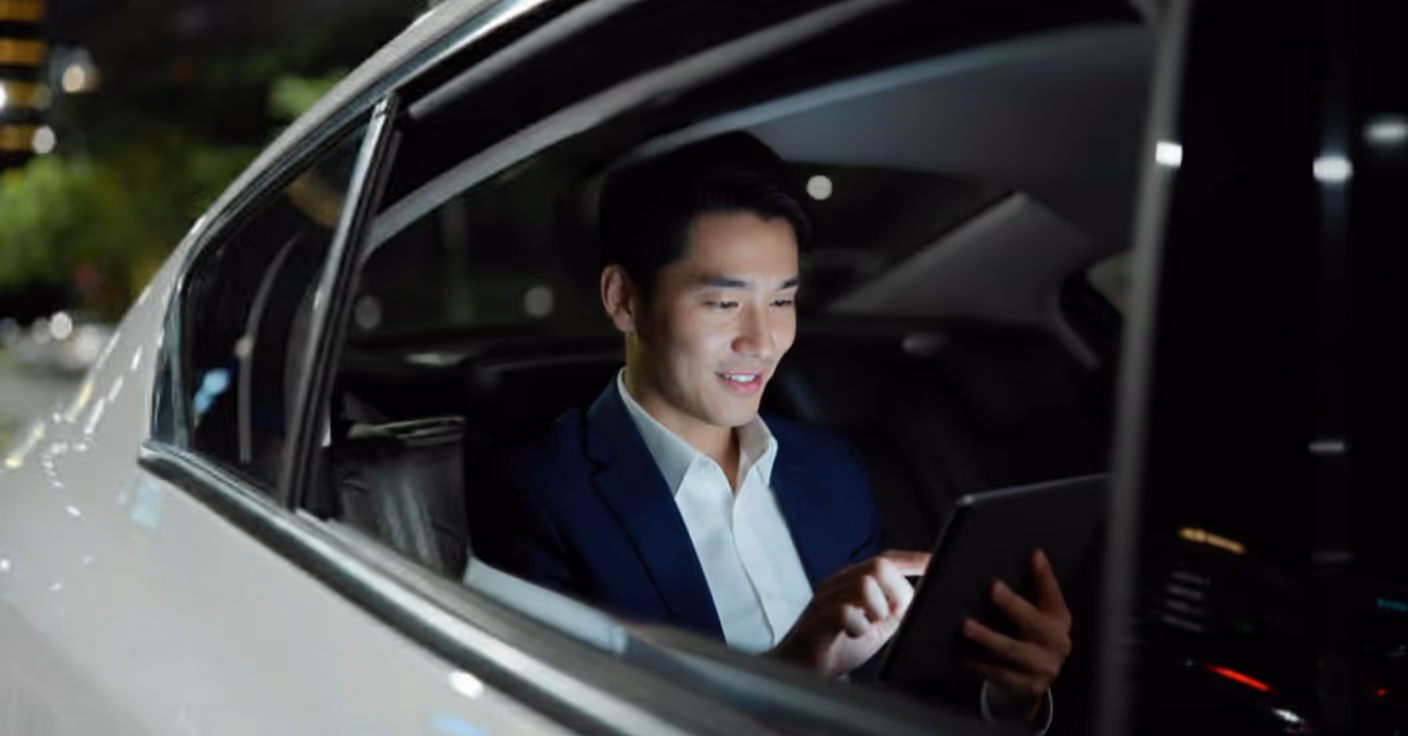 Un homme en costume assis à l'arrière d'une voiture la nuit, utilisant une tablette avec un sourire.
