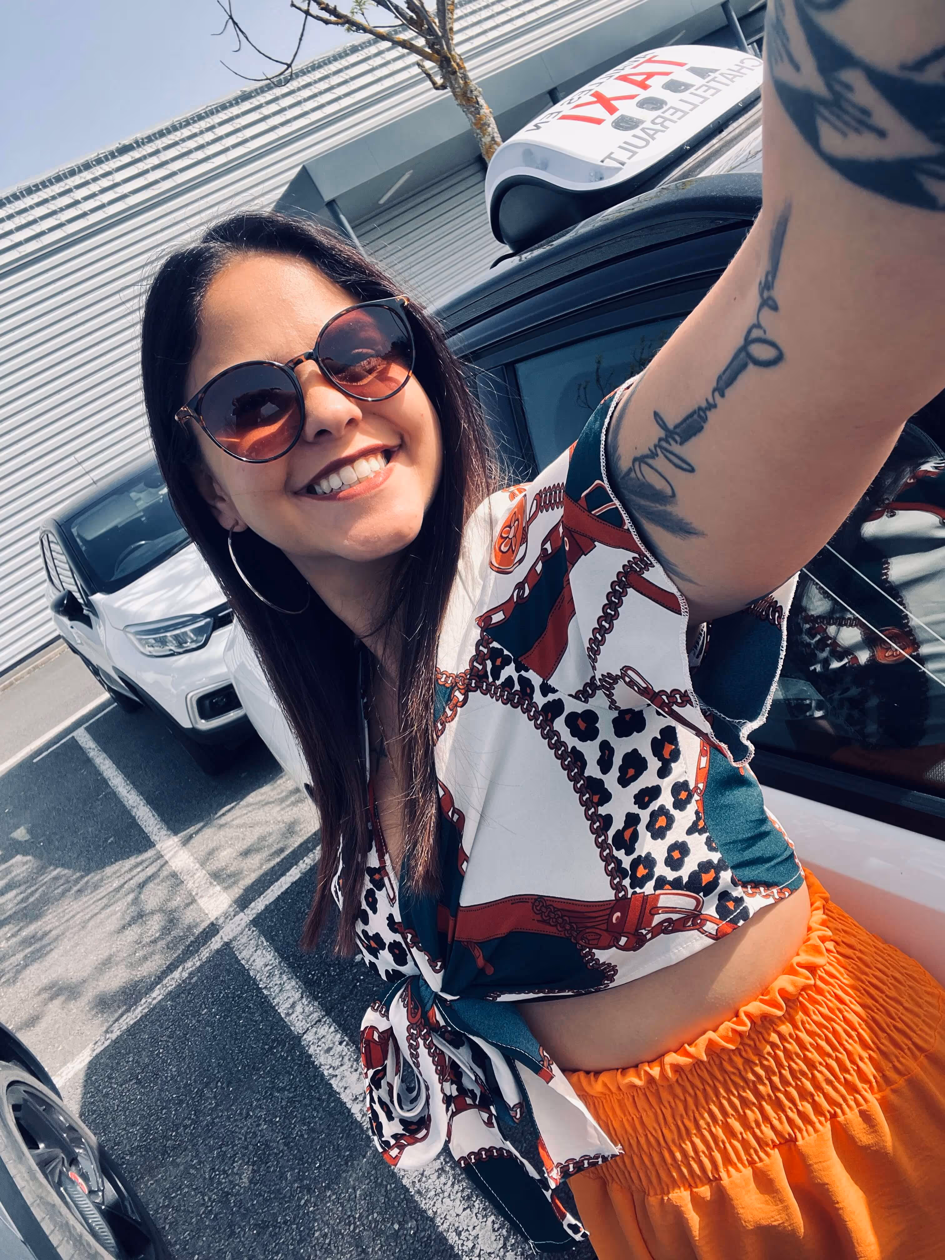 Femme souriante portant des lunettes de soleil et un haut coloré prenant un selfie en plein air, debout près d'une voiture de taxi.