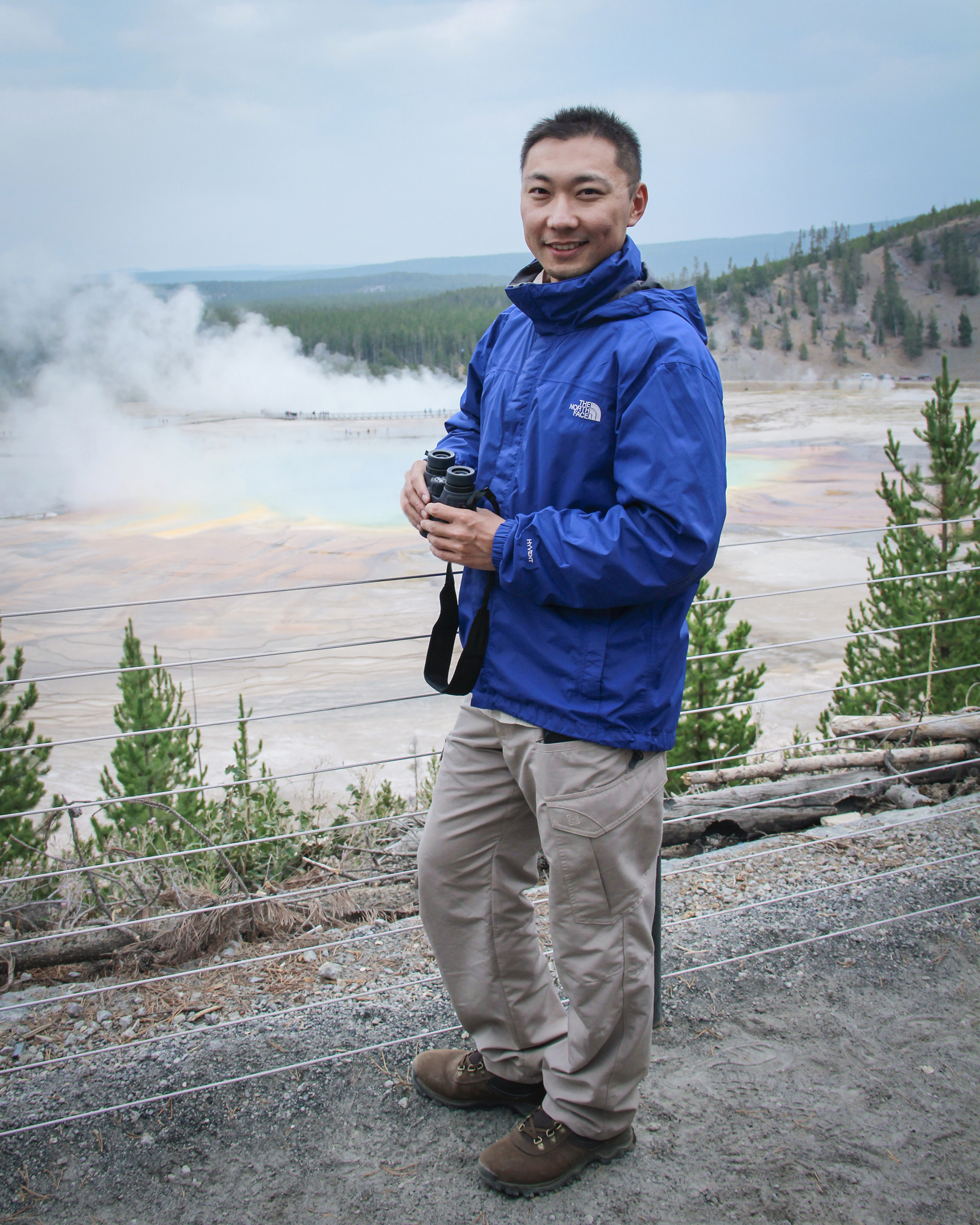 Smiling man in a blue jacket holding binoculars standing near a colorful steaming geothermal pool surrounded by trees.