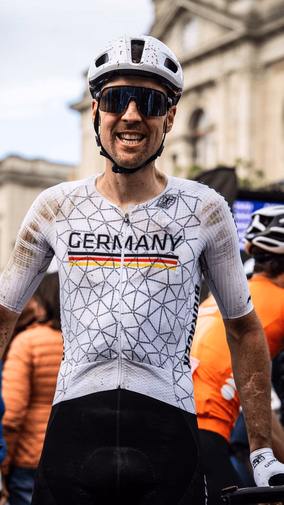Schmutziger Radfahrer mit weißem Helm und Deutschland-Trikot lächelt nach einem Rennen.