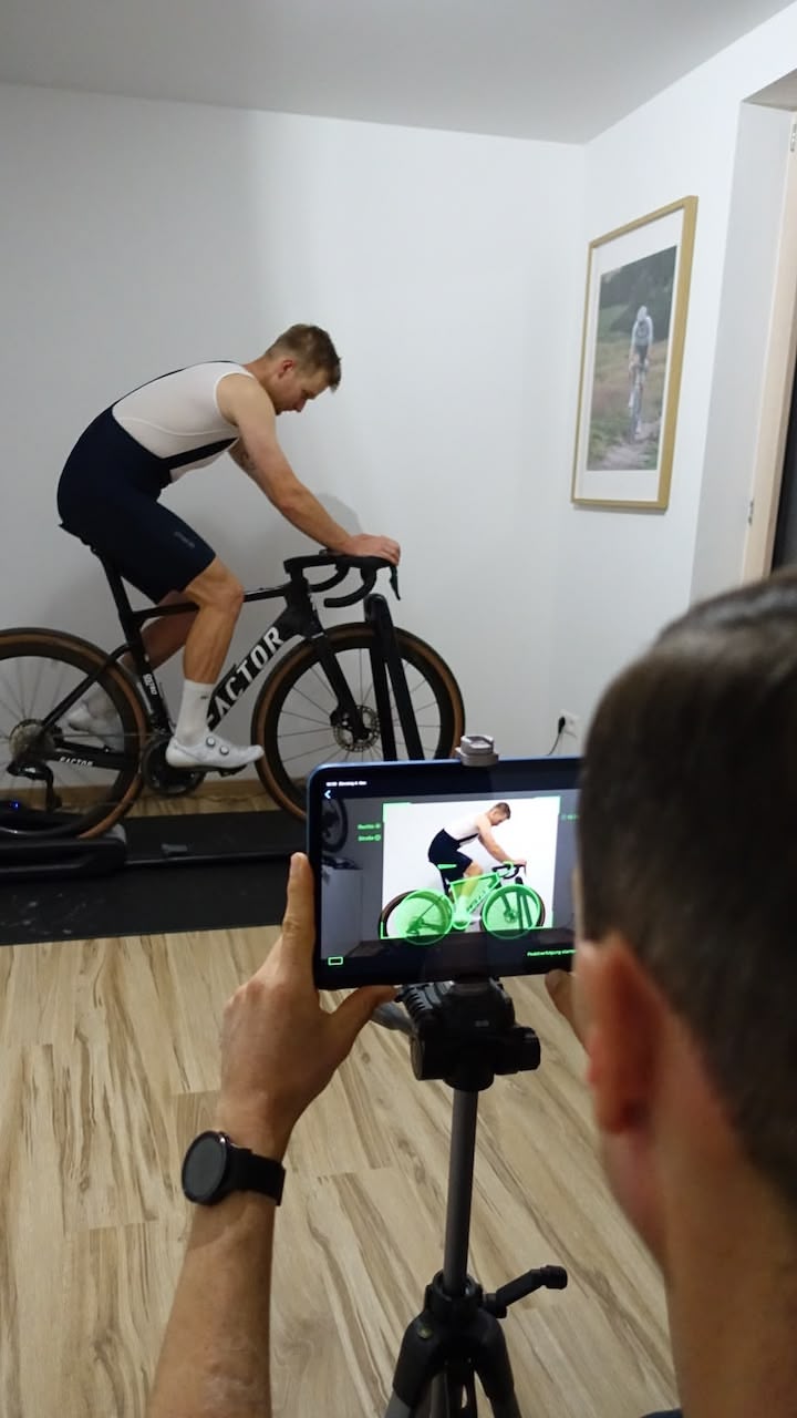 Ein Mann auf einem Fahrrad im Training auf einem Rollentrainer, während eine andere Person mit einem Tablet sein Fahrbild aufnimmt.