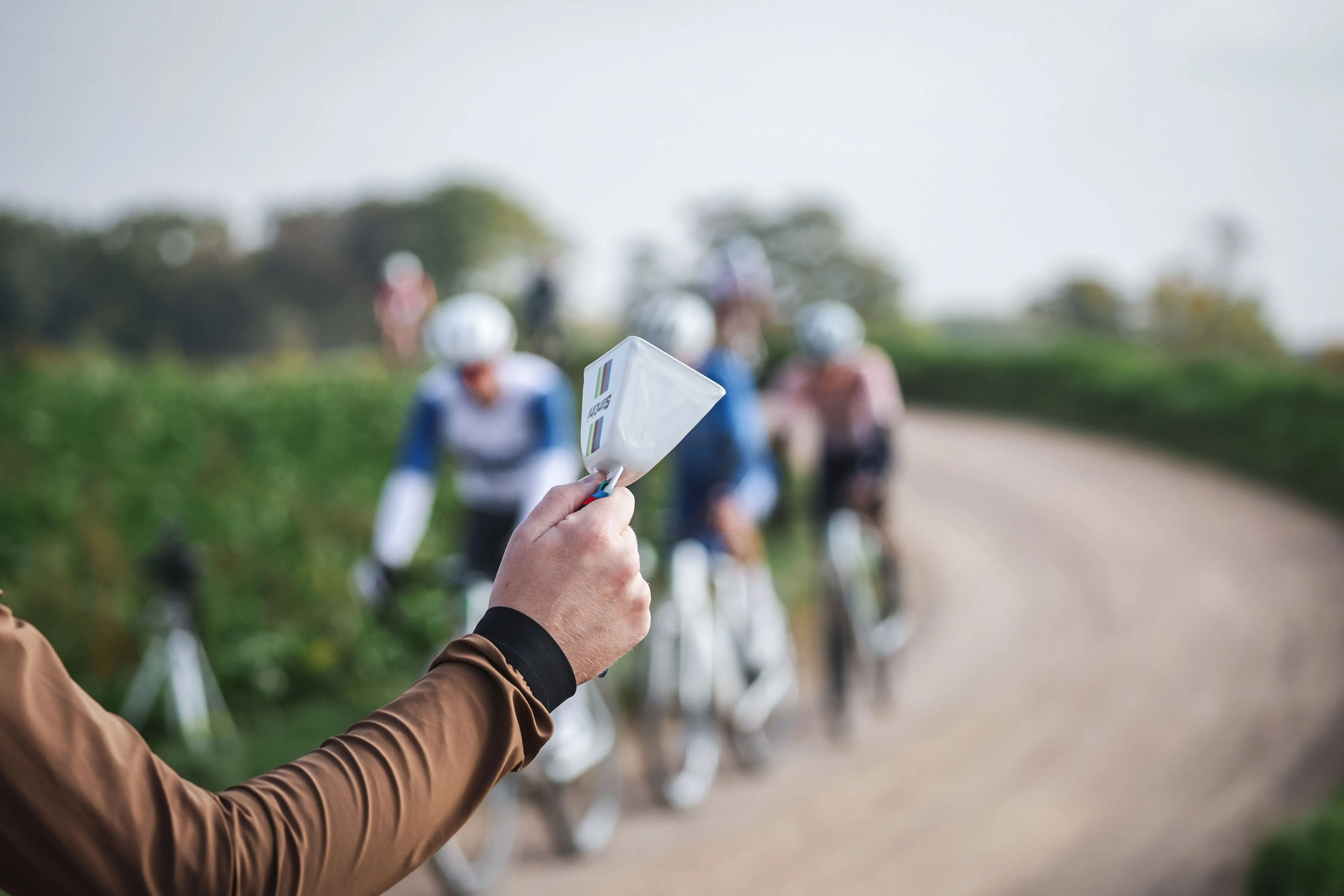 Hand hält weiße Glocke mit Aufschrift "WOLLINGER" vor einer Gruppe Radfahrer auf einem gewundenen Feldweg.