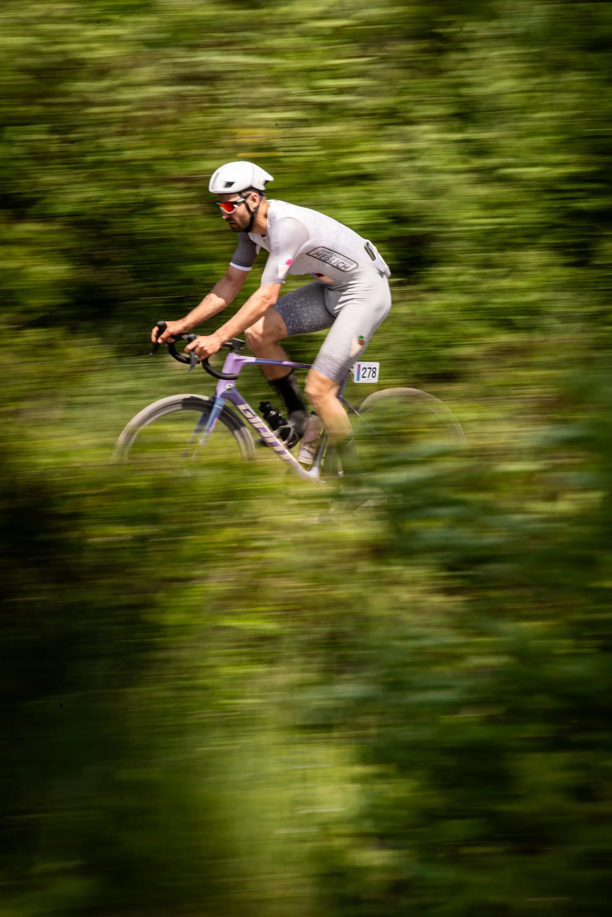 Radsportler in grauer Kleidung und Helm fährt mit Rennrad schnell durch bewaldetes Gebiet.