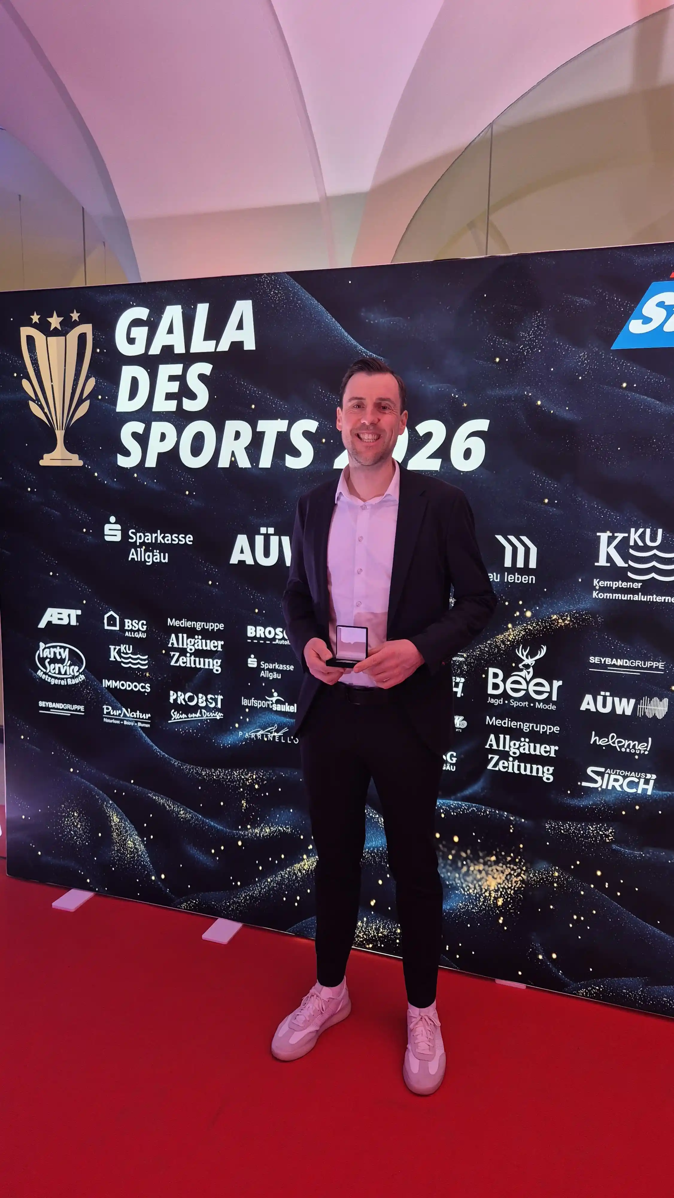 Ein lächelnder Mann in einem dunklen Anzug und weißen Sneakern steht auf einem roten Teppich vor einer Wand mit der Aufschrift „Gala des Sports 2026“ und mehreren Sponsorenlogos, er hält eine Auszeichnung in den Händen.