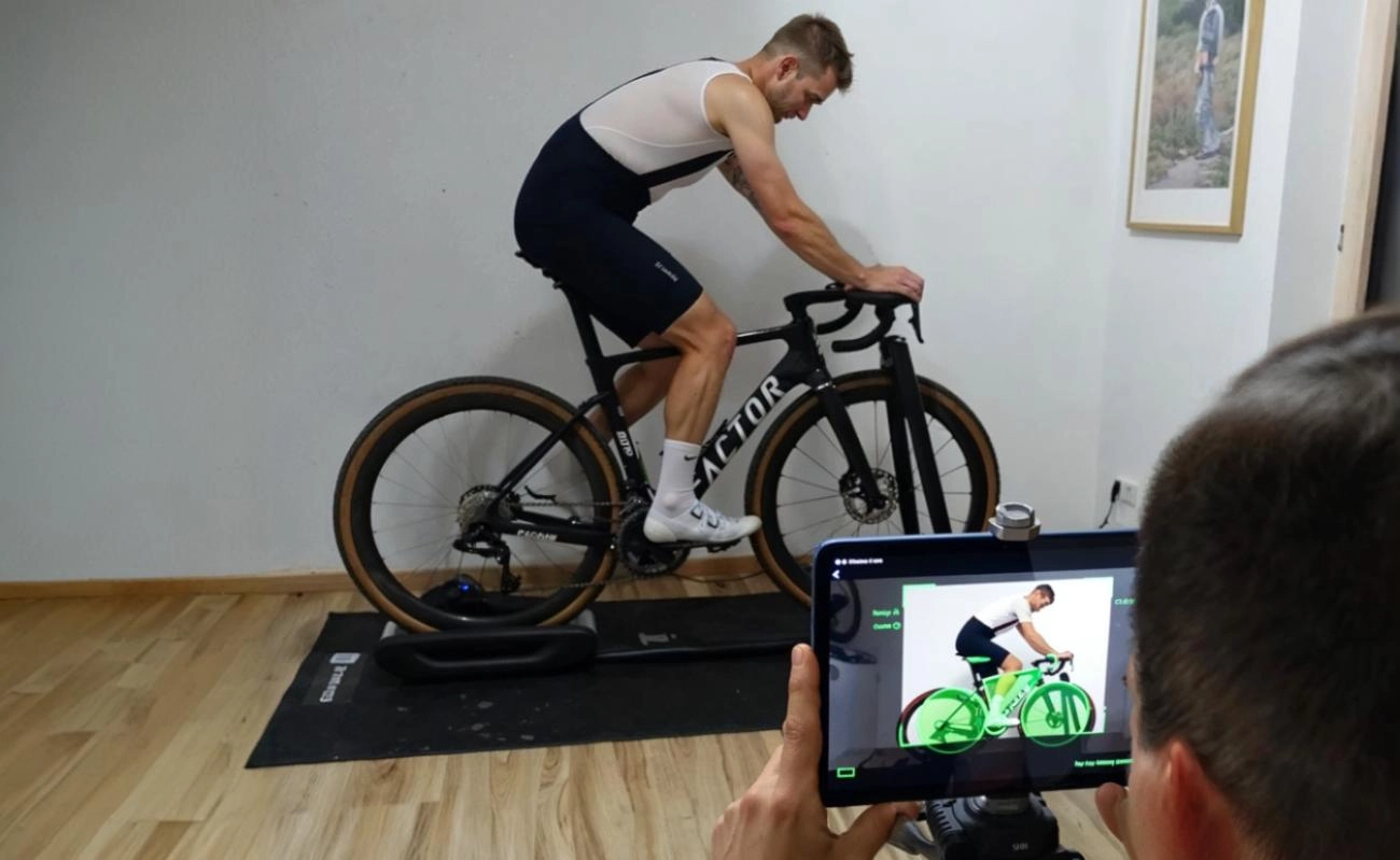 Ein Mann in Radsportkleidung sitzt auf einem Fahrrad in einem Raum, während eine andere Person eine Tablet-Kamera verwendet, um ihn für eine Fahrradvermessung zu filmen.