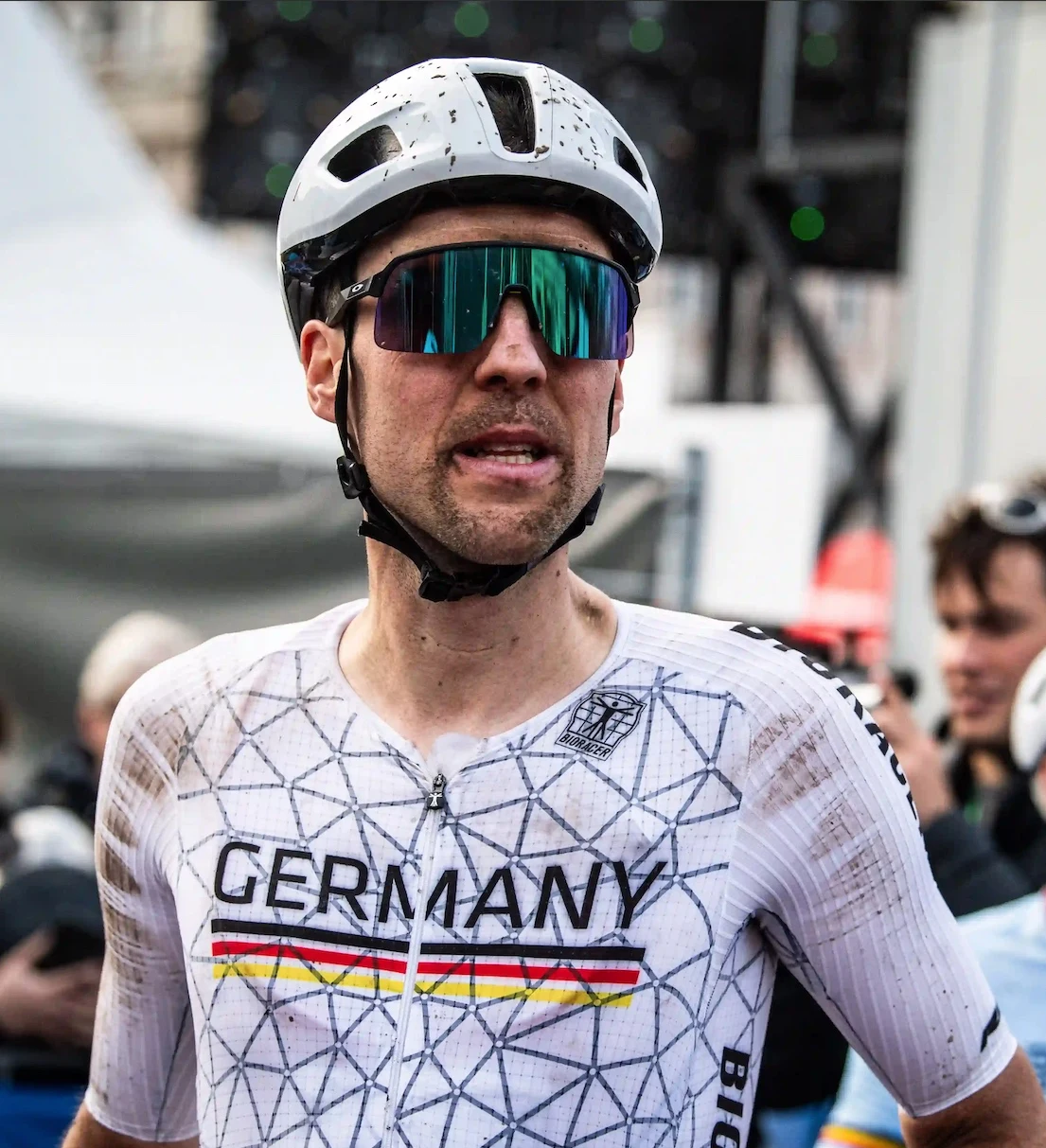 Radfahrer in weißem Trikot mit der Aufschrift „GERMANY“ und dreifarbigen Streifen, Helm und reflektierender Sonnenbrille, mit Schmutzflecken übersät.