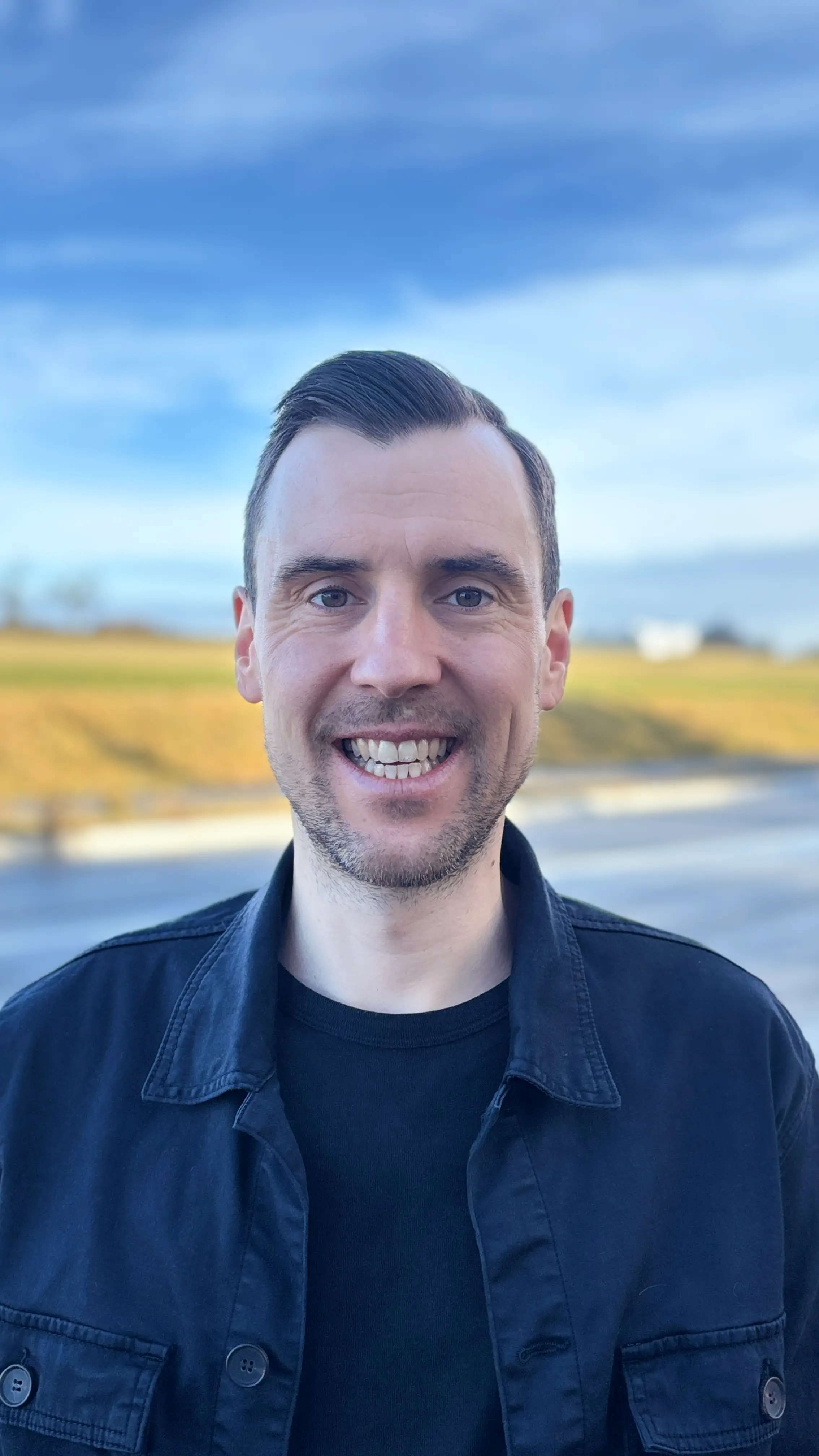 Lächelnder Mann mit kurzem Haar, schwarzem T-Shirt und schwarzer Jacke vor einem unscharfen Hintergrund aus blauem Himmel und grüner Landschaft.