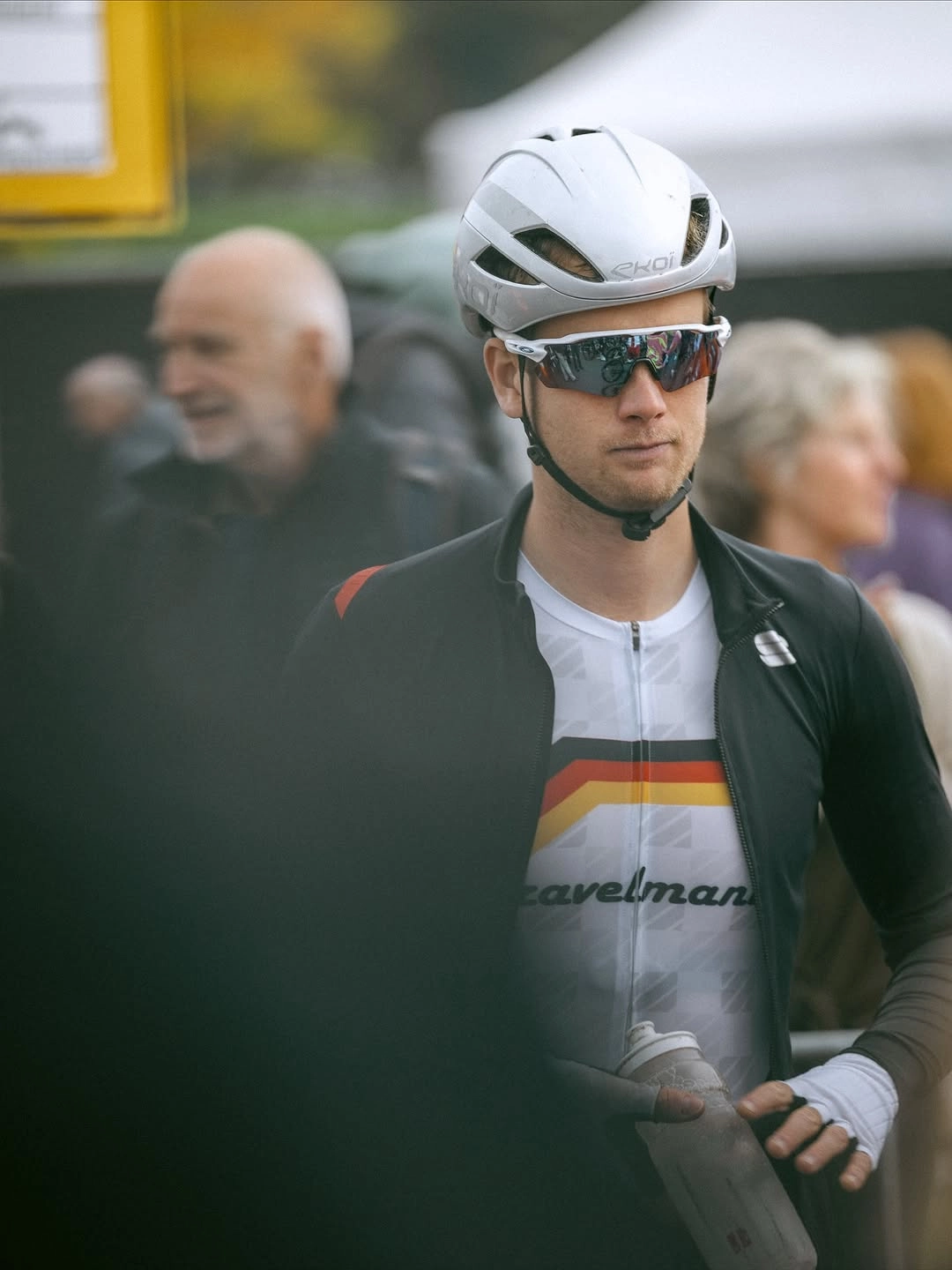Junger Radfahrer mit weißem Helm und Sonnenbrille hält eine Trinkflasche, unscharfe Menschen im Hintergrund.