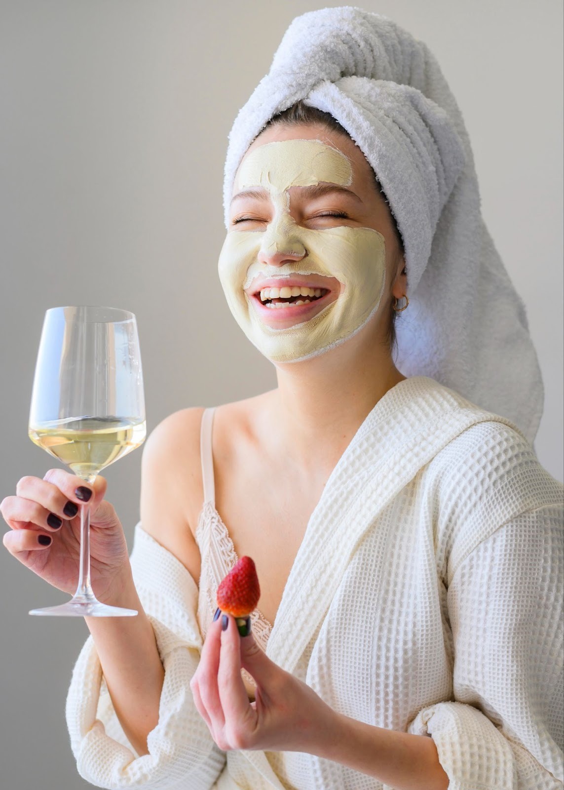 A girl in bathrobe, and hair wrapped in towel has applied facemask, is laughing with a wine glass in one hand and a strawberry in other.
