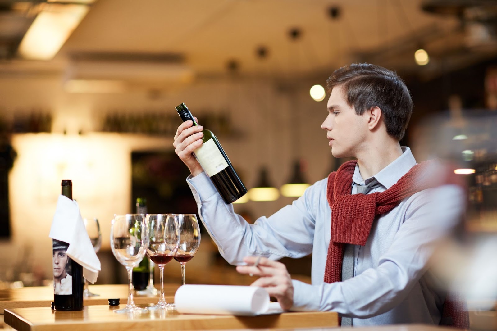 A man sitting besides a table where a bottle of wine and 3 glasses are kept with a notepad. He is looking at the label of a wine bottle in his one hand and has pen in other.