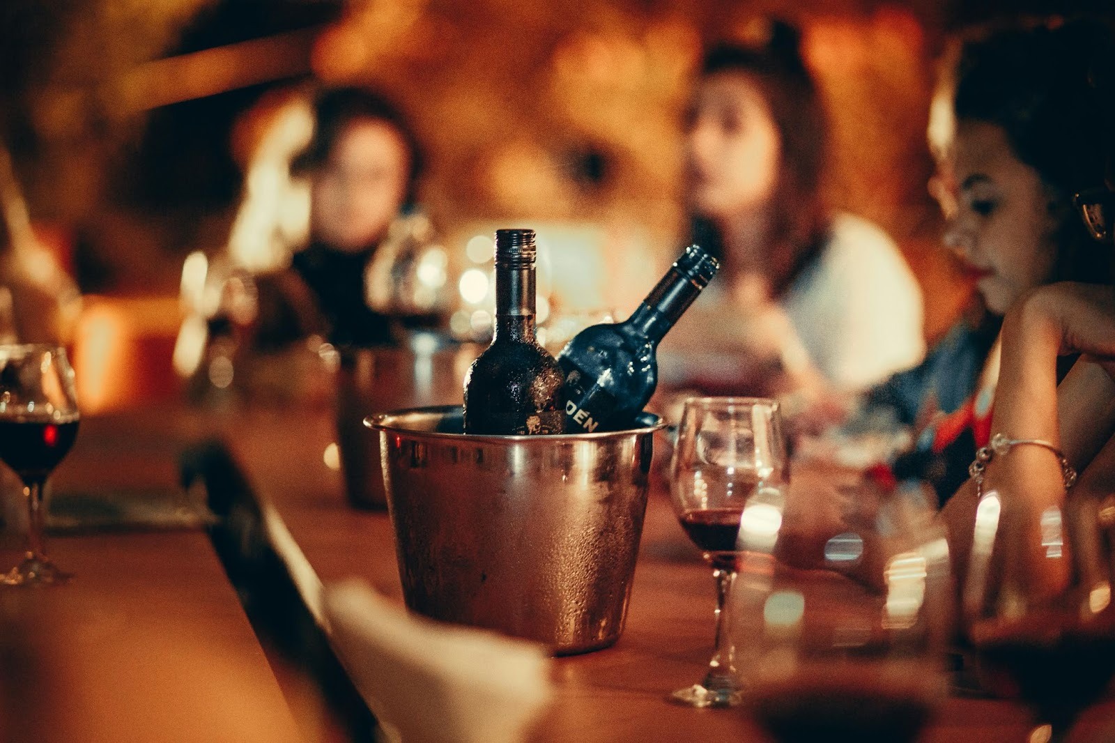 Two wine bottles in ice bucket and wine glasses filled on a table surrounded by ladies 