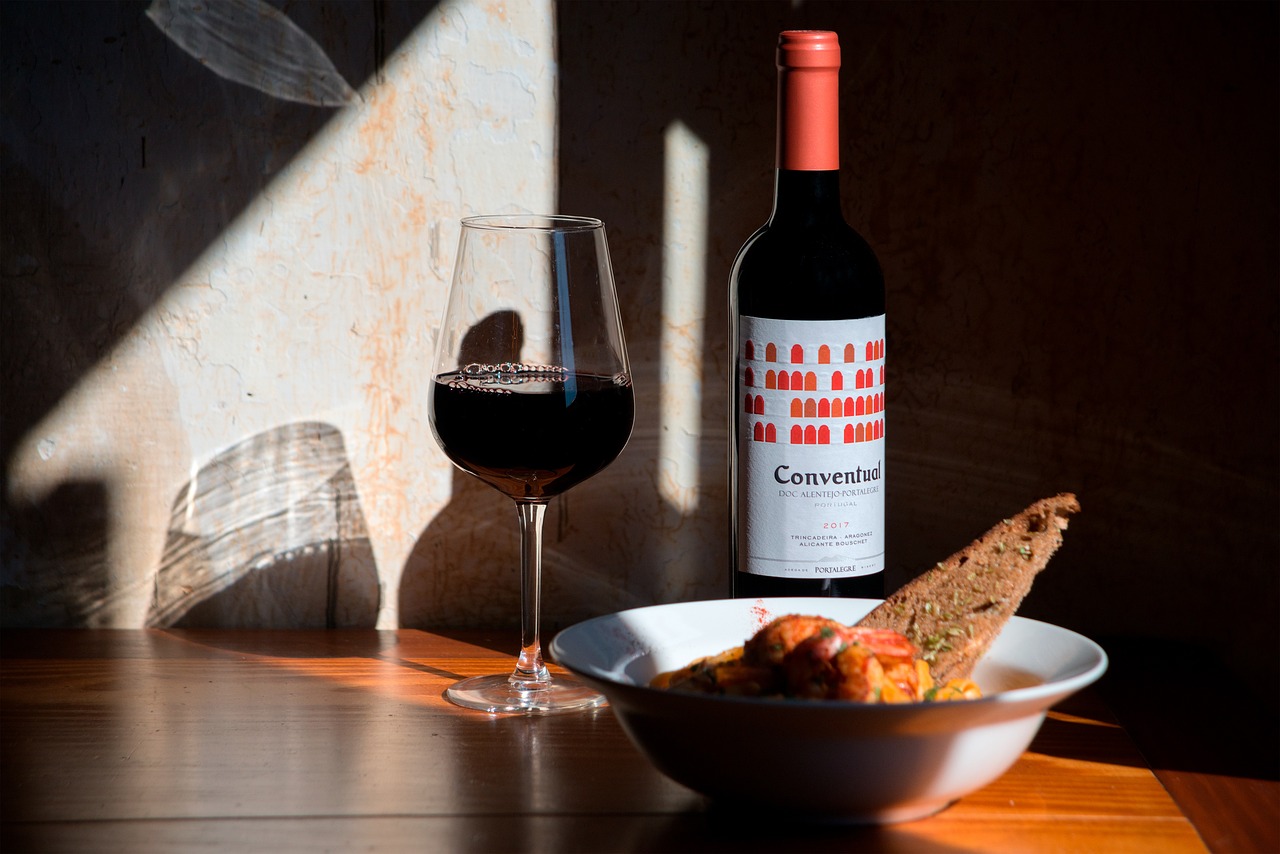 A red wine bottle , a filled glass and a bowl of food kept on a wooden surface and light hitting from one side