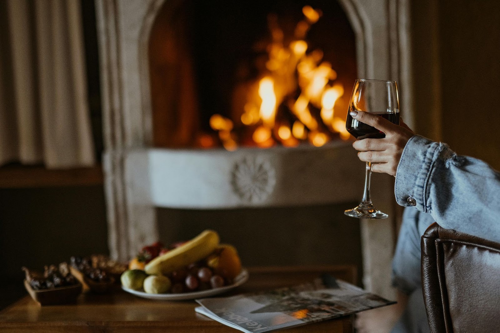 A person sitting in front of a fire place, corner of chair and the hand holding wine in view next to a table where a plate of fruits and a magazine is kept