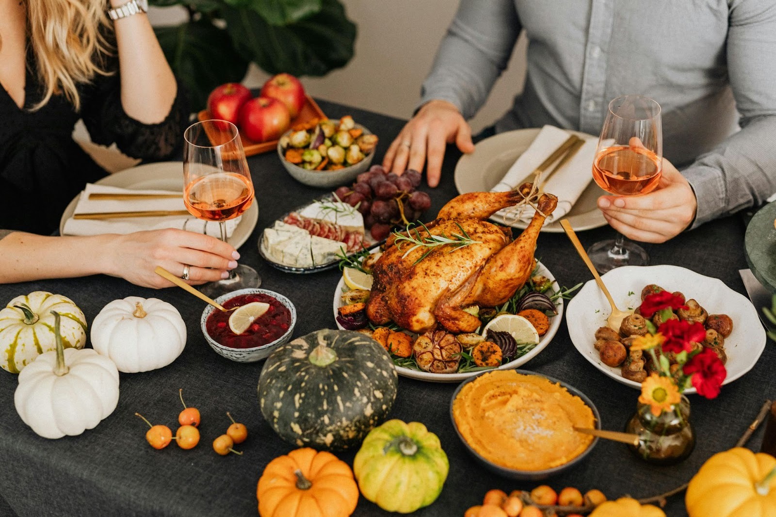 two people sitting around a table full of food items, holding a glass of wine each