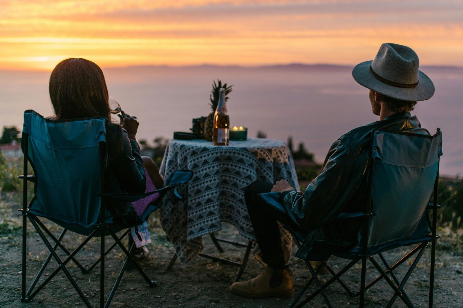 A couple sitting on chairs around a table having a wine bottle, a pineapple and candles on it. They are looking at the sunset on the mountains. 