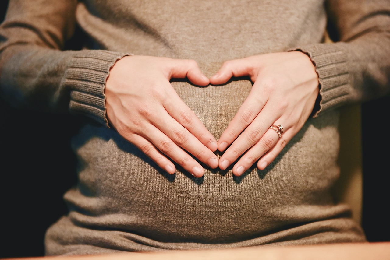 A pregnant woman wearing brown sweater has kept her hands on her belly making a heart shape