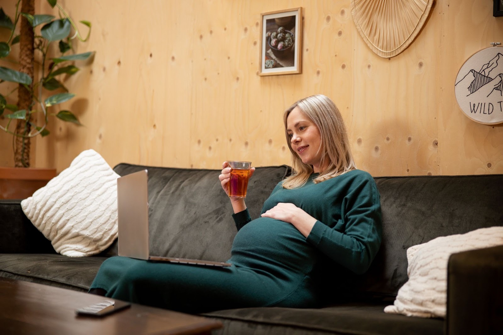 A pregnant woman sitting on a couch, laptop on her lap and an orange drink glass in her hand inside a wooden room