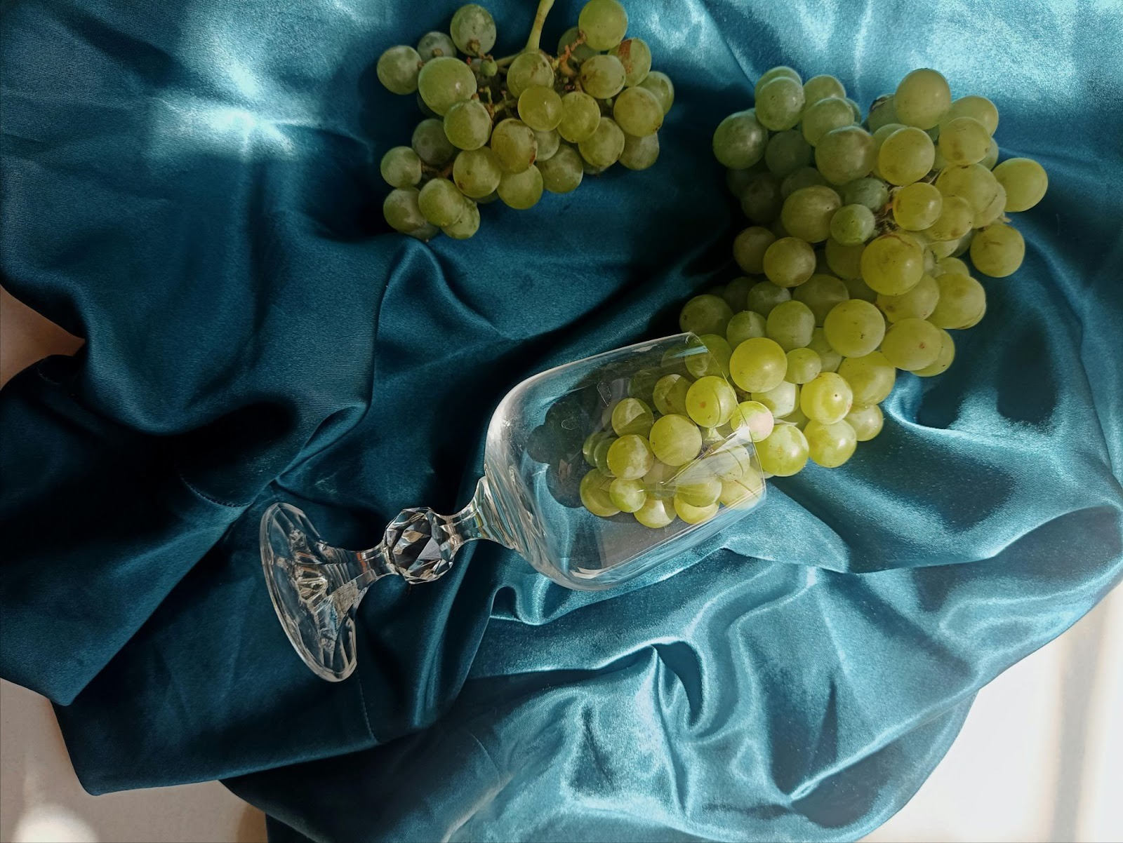 A wine glass tilted a bunch of grapes some inside and some out laying near it on a blue silk cloth