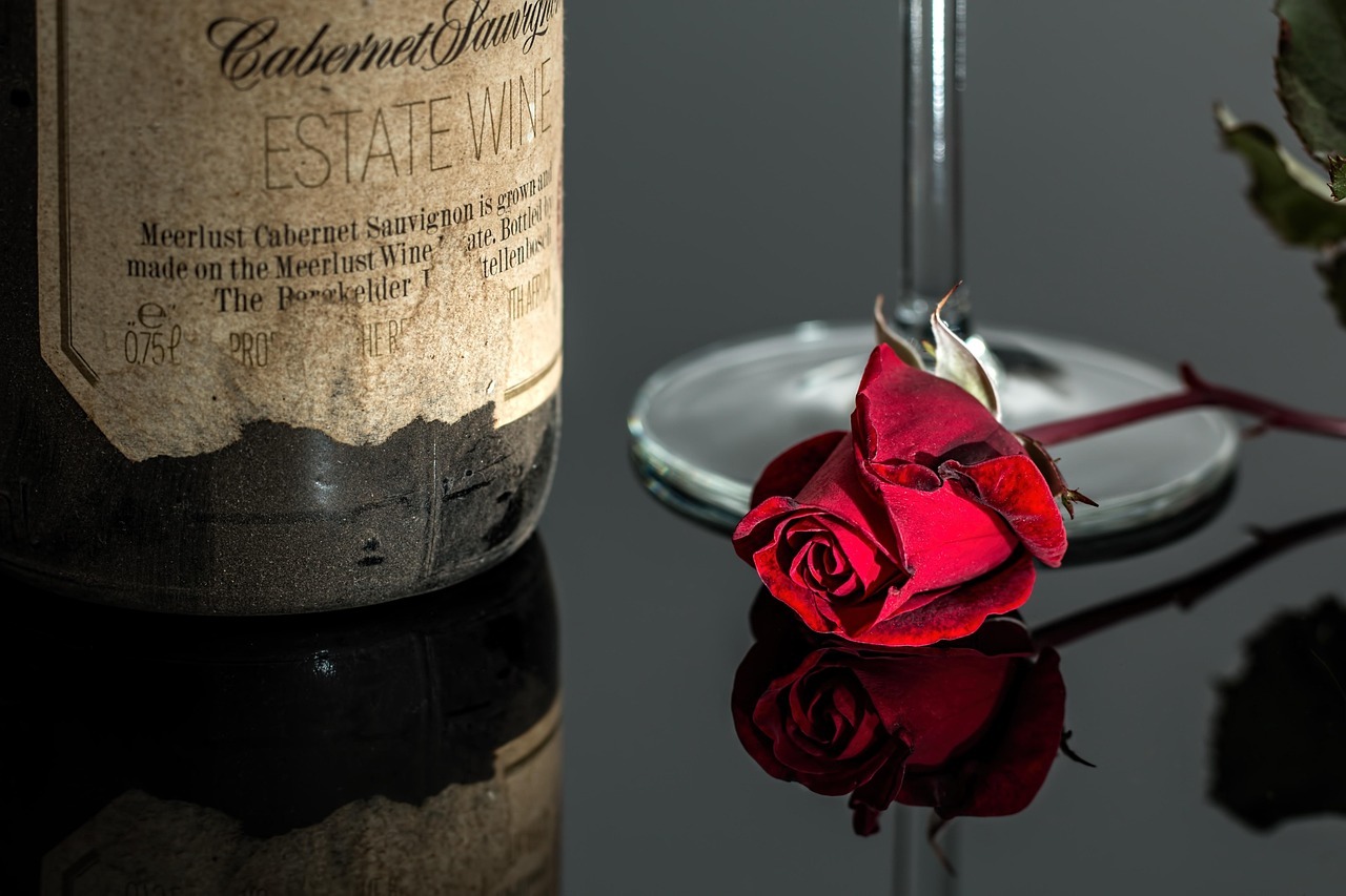 Lower part of a wine bottle and a wine glass visible on a glass surface with a red rose in front.