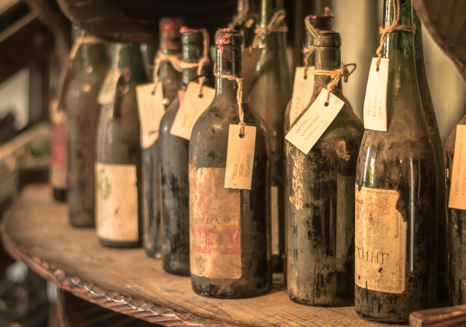 multiple old wine bottles on a wooden shelf with worn out labels and dust on them