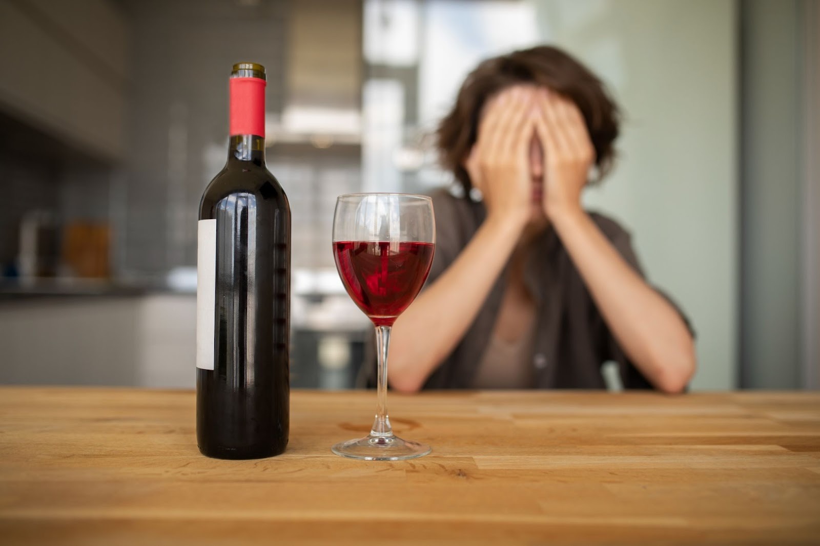 a girl with face covered with her hands, sitting in her kitchen, a wine bottle and a filled glass kept in front on a wooden surface.