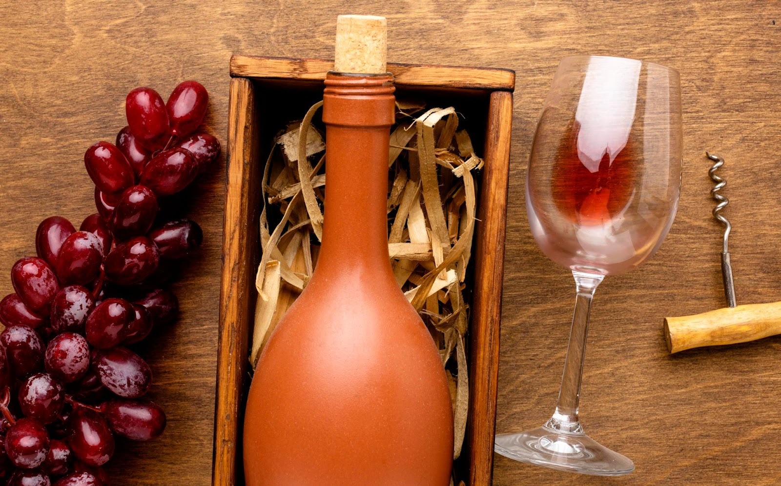 An orange wine bottle in a wooden box next to red grapes on one side and a wine glass and cork screw on other