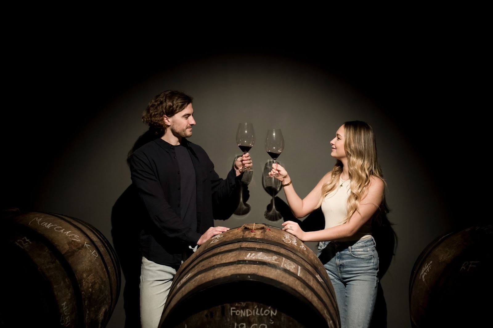 A man and a woman standing on opposite sides of a barrel holding wine glass in their hand facing each other