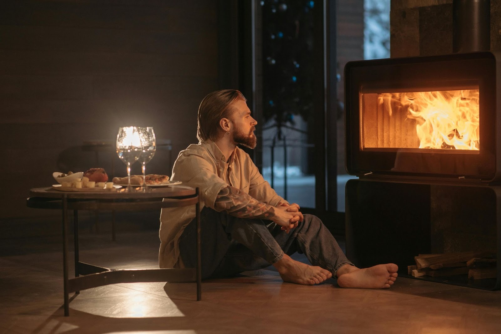 A man sitting on floor by the fireplace and some food and 2 glasses of wine kept on the table nearby