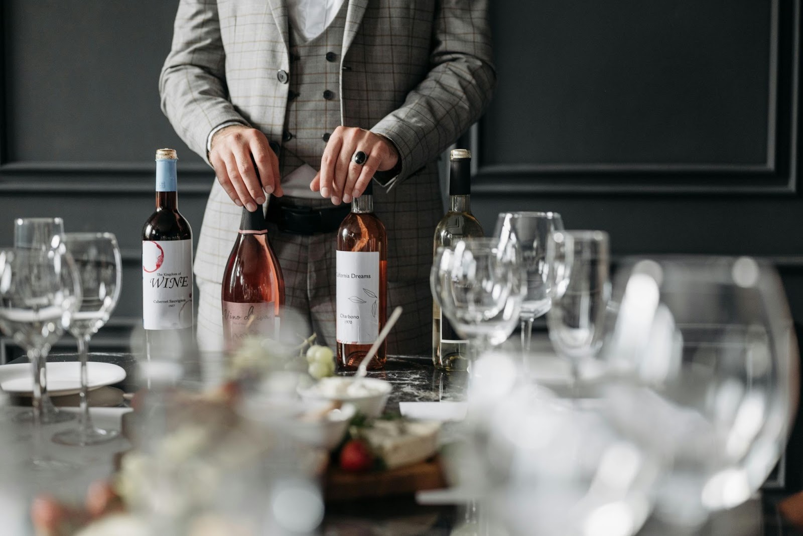 A man standing next to a table containing lot of wine glasses and four wine bottles and he has his hands on two bottles
