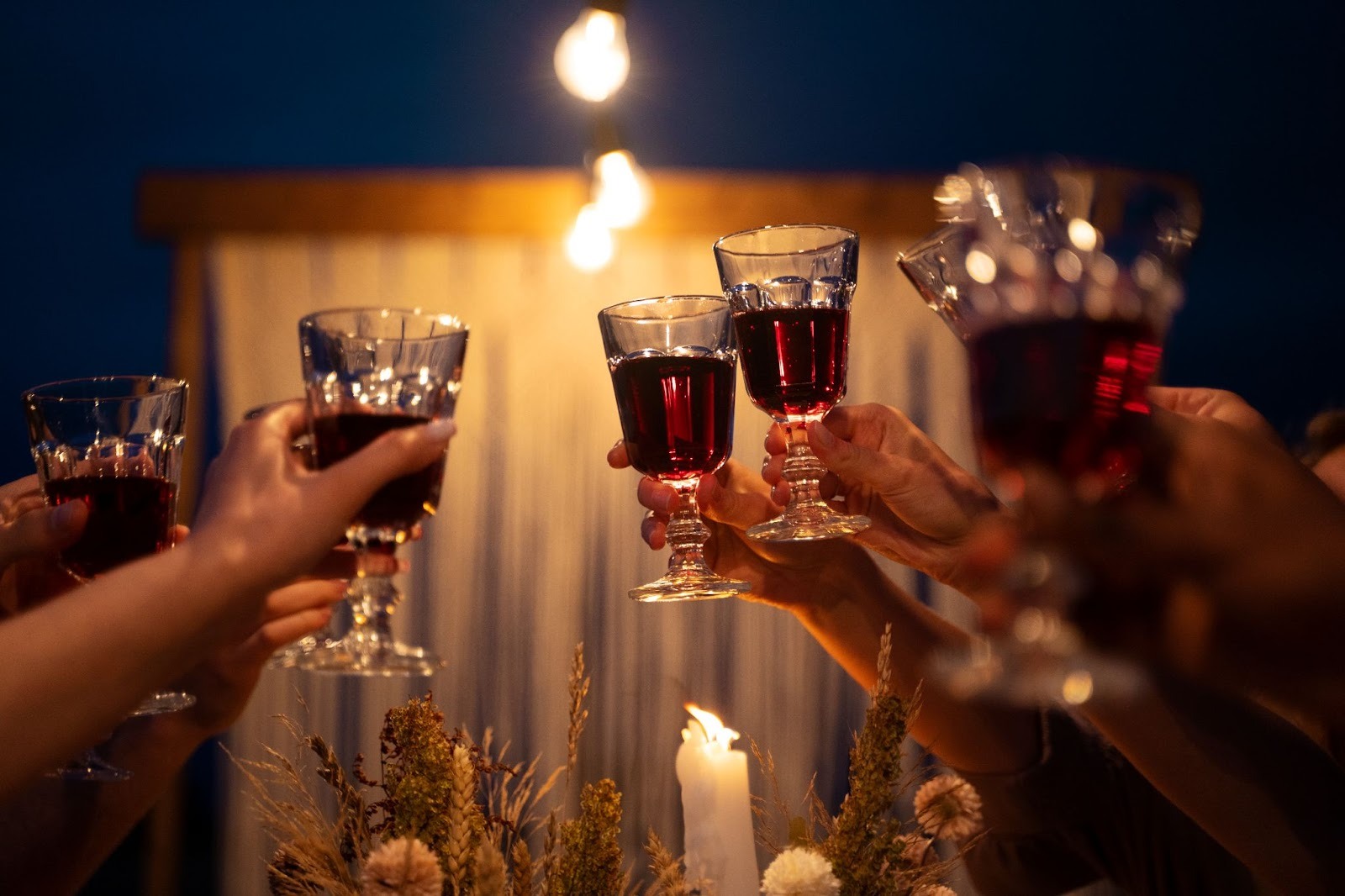multiple hands holding wine glasses with some dried flowers and a candle beneath 