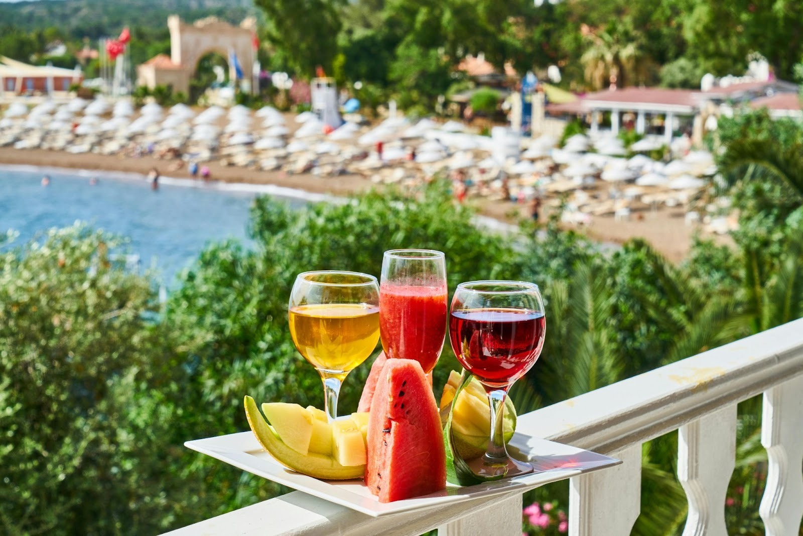 three wine glasses and some fruits in a plate on the railing of a balcony and beach area in background