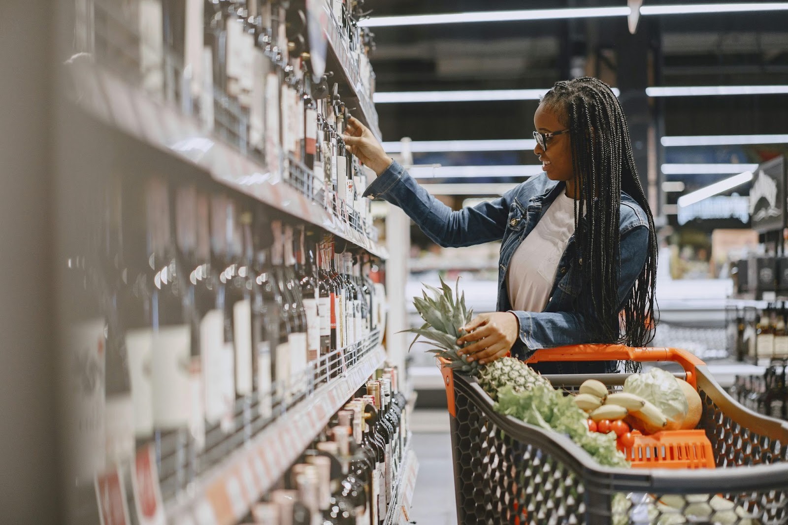 A girl with shopping cart looking at wine bottles in mart. 