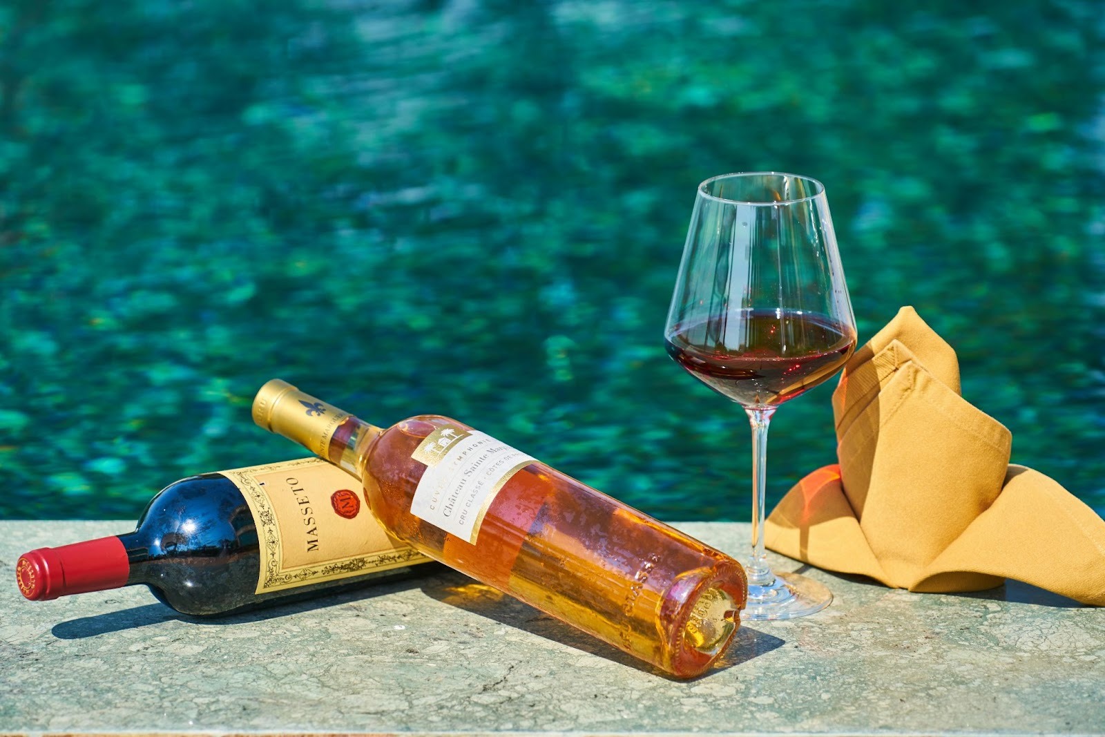 two wine bottles laying near waterbody with a wine glass and cloth napkin nearby