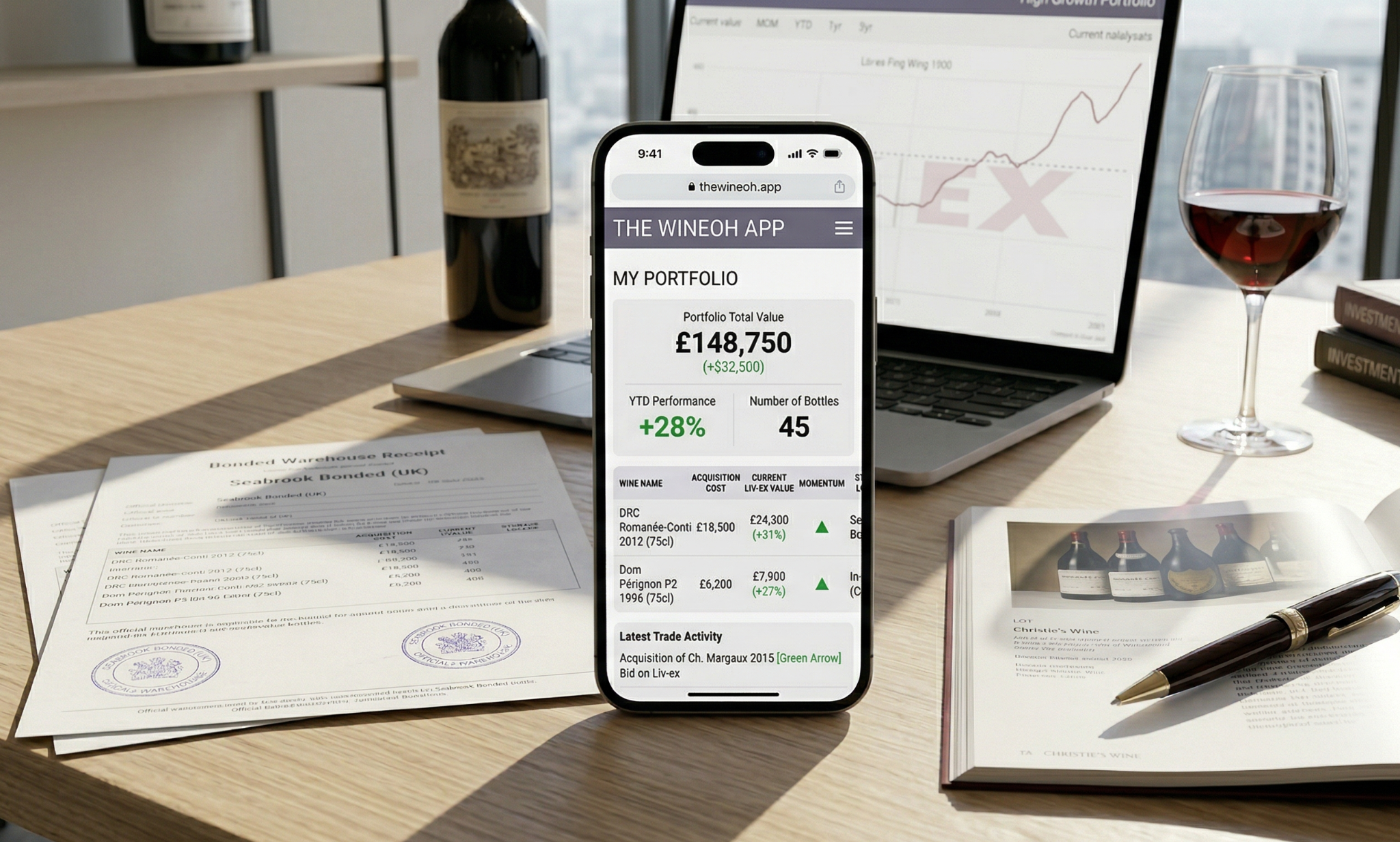 A laptop, wine bottle, a glass, some papers, a magazine with pen and a phone with the wine oh app open on it kept on a table