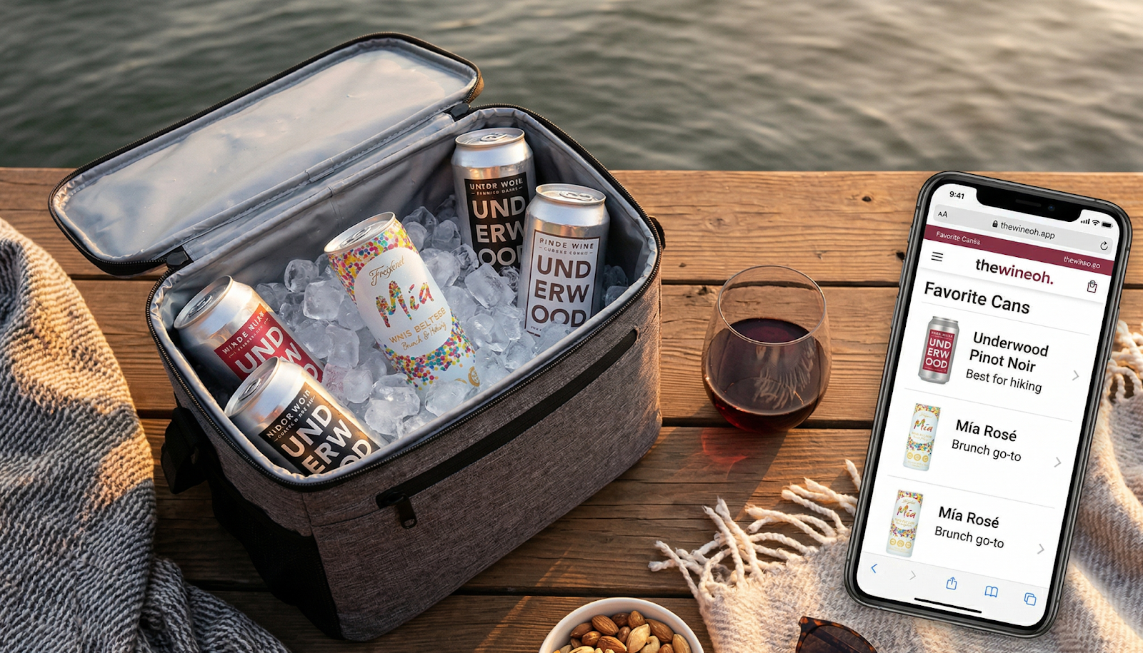 A cooler filled with ice and canned wines, a wine glass resting beside it half-filled, and a smartphone displaying thewineoh.app with a Favorite Cans list