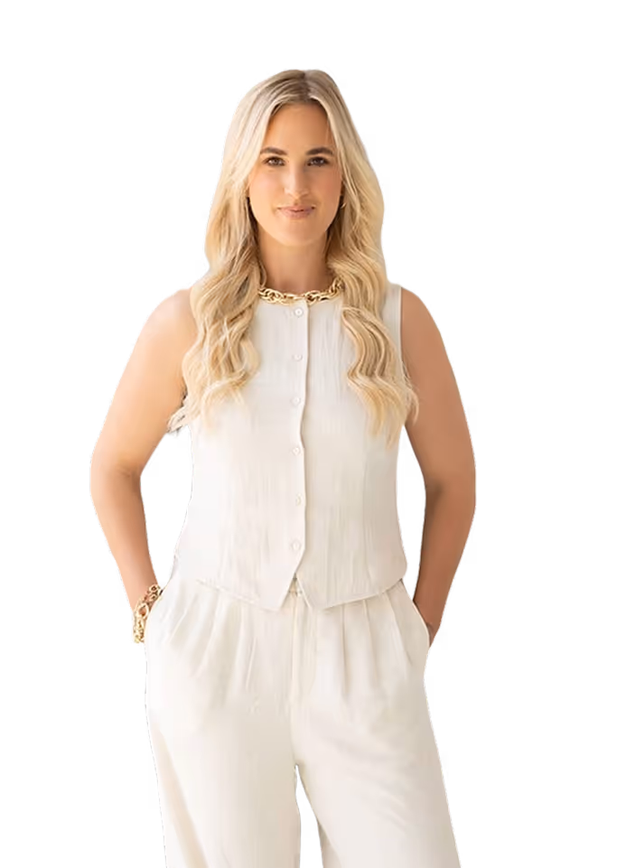 Blonde woman with long wavy hair wearing a sleeveless white buttoned top and white pants, standing with hands in pockets.