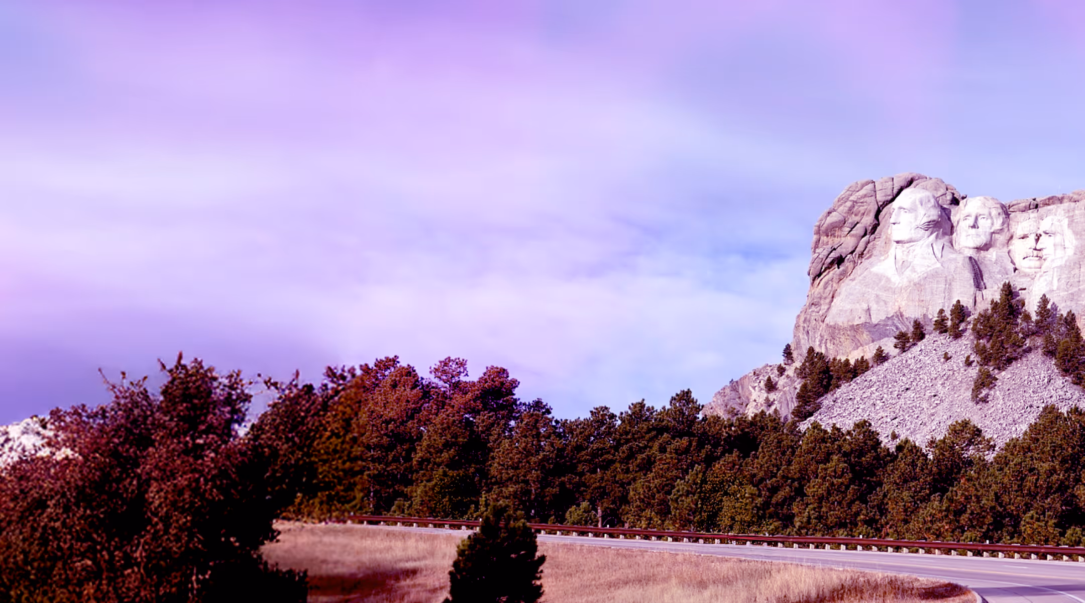 Mount Rushmore with carved faces of U.S. presidents overlooking a forest and curved road under a purple-tinged sky.