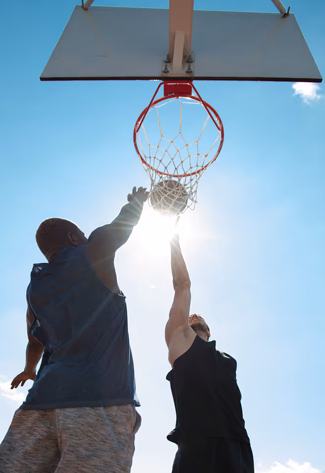 Two men playing basketball outdoors as one attempts a shot near the hoop with the sun shining behind the net.