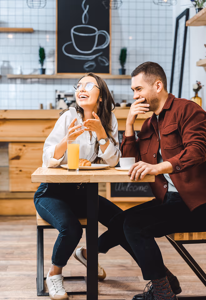 Smiling young man and woman sitting at a café table with a croissant, orange juice, and coffee, enjoying a conversation.