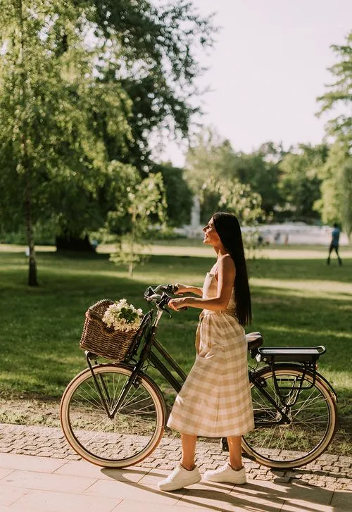 Woman in a checkered skirt standing beside a bicycle with a basket of flowers in a sunlit park.