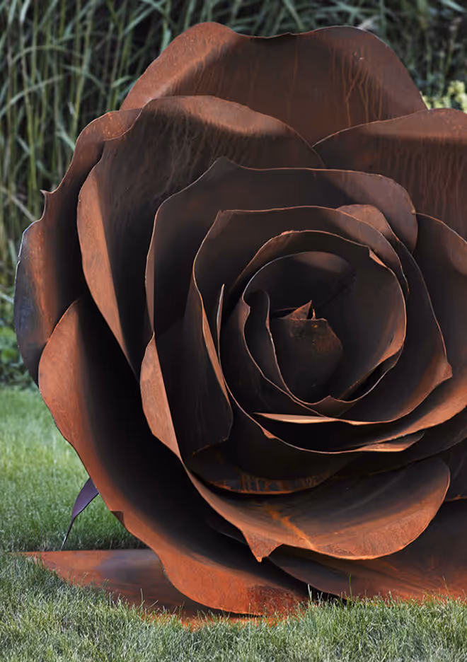 Large rust-colored metal sculpture shaped like a rose placed on grass with tall greenery in the background.