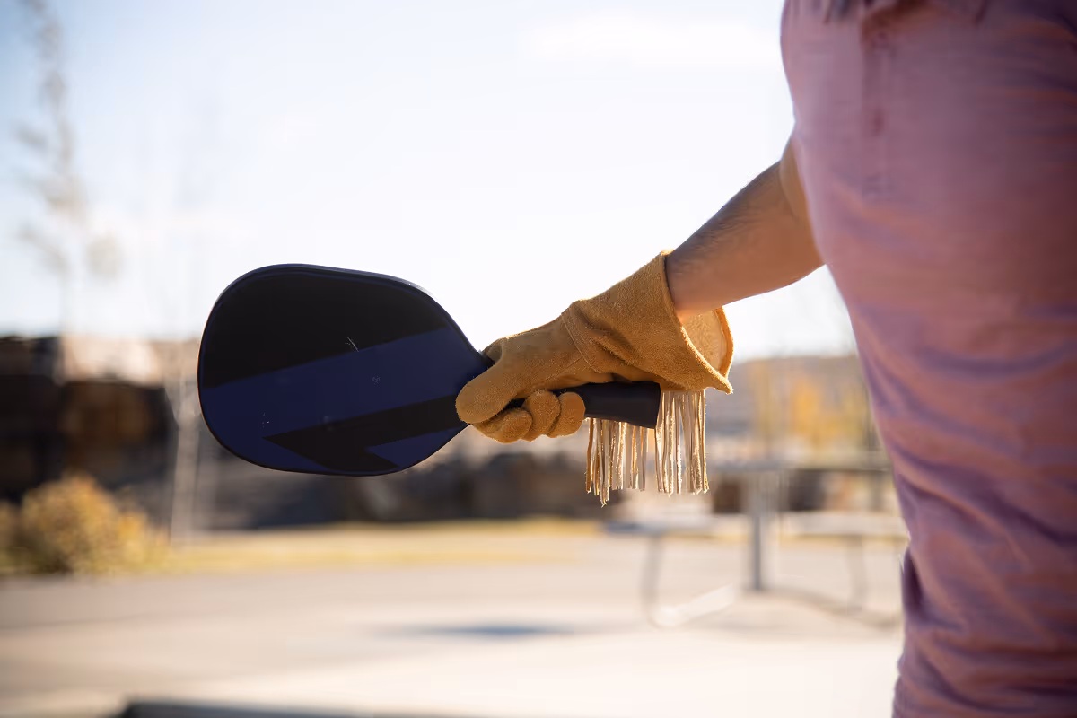Person wearing a glove holding a black pickleball paddle outdoors with blurred background.