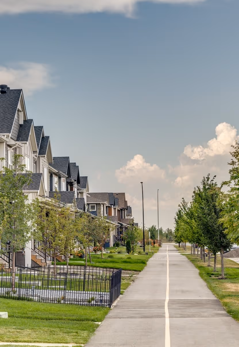 Residential street with rows of houses on the left and a paved walking path lined with trees on the right under a partly cloudy sky.