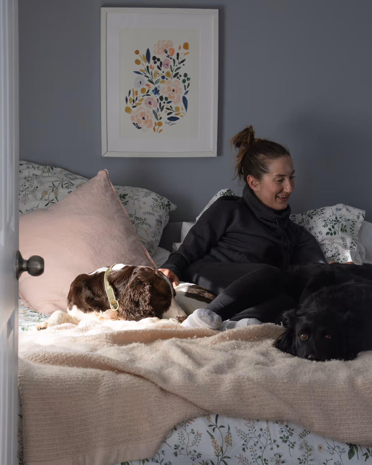 Woman sitting on a bed with a brown and white dog and a black dog resting beside her.