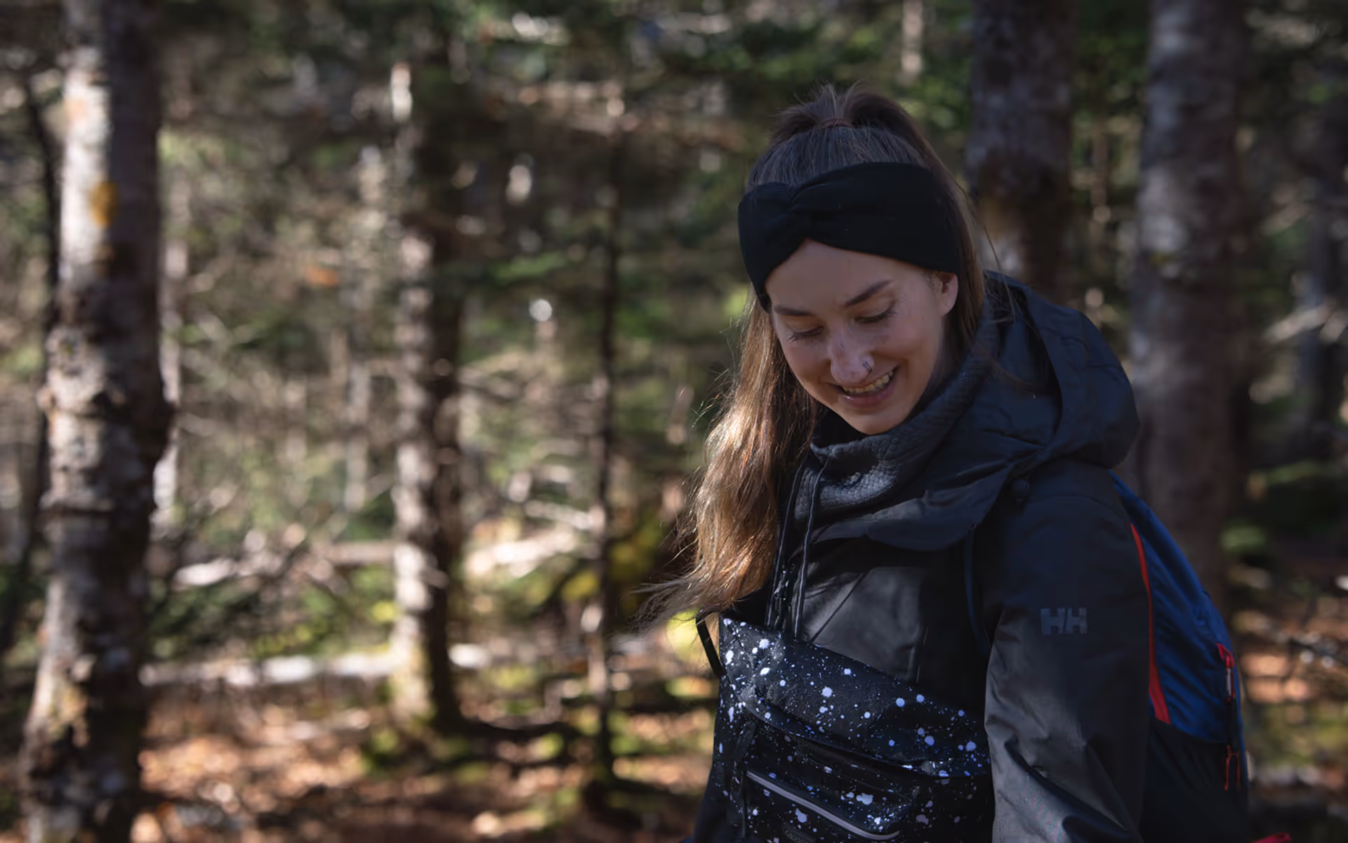 Smiling woman wearing a black headband and jacket standing in a sunlit forest.