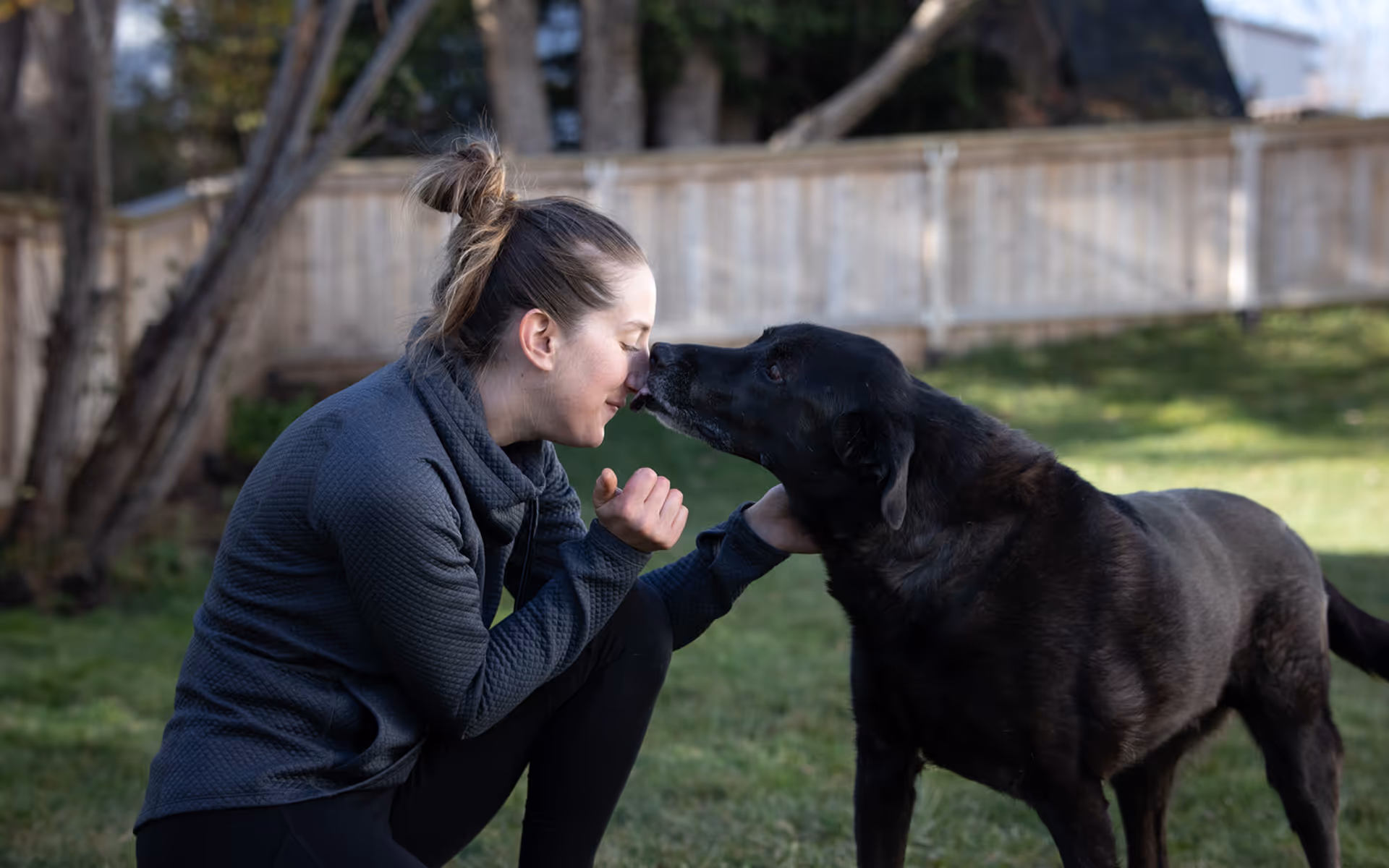 Woman with hair in a bun wearing a gray sweater kneeling on grass, touching noses with a large black dog in a backyard.