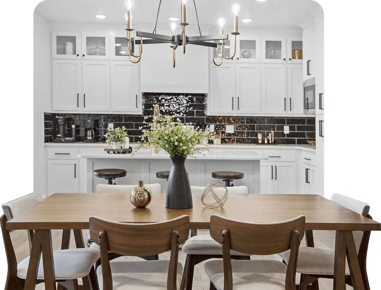 Modern dining area with wooden table, six upholstered chairs, black vase with green flowers, and a kitchen with white cabinets and black backsplash in the background.